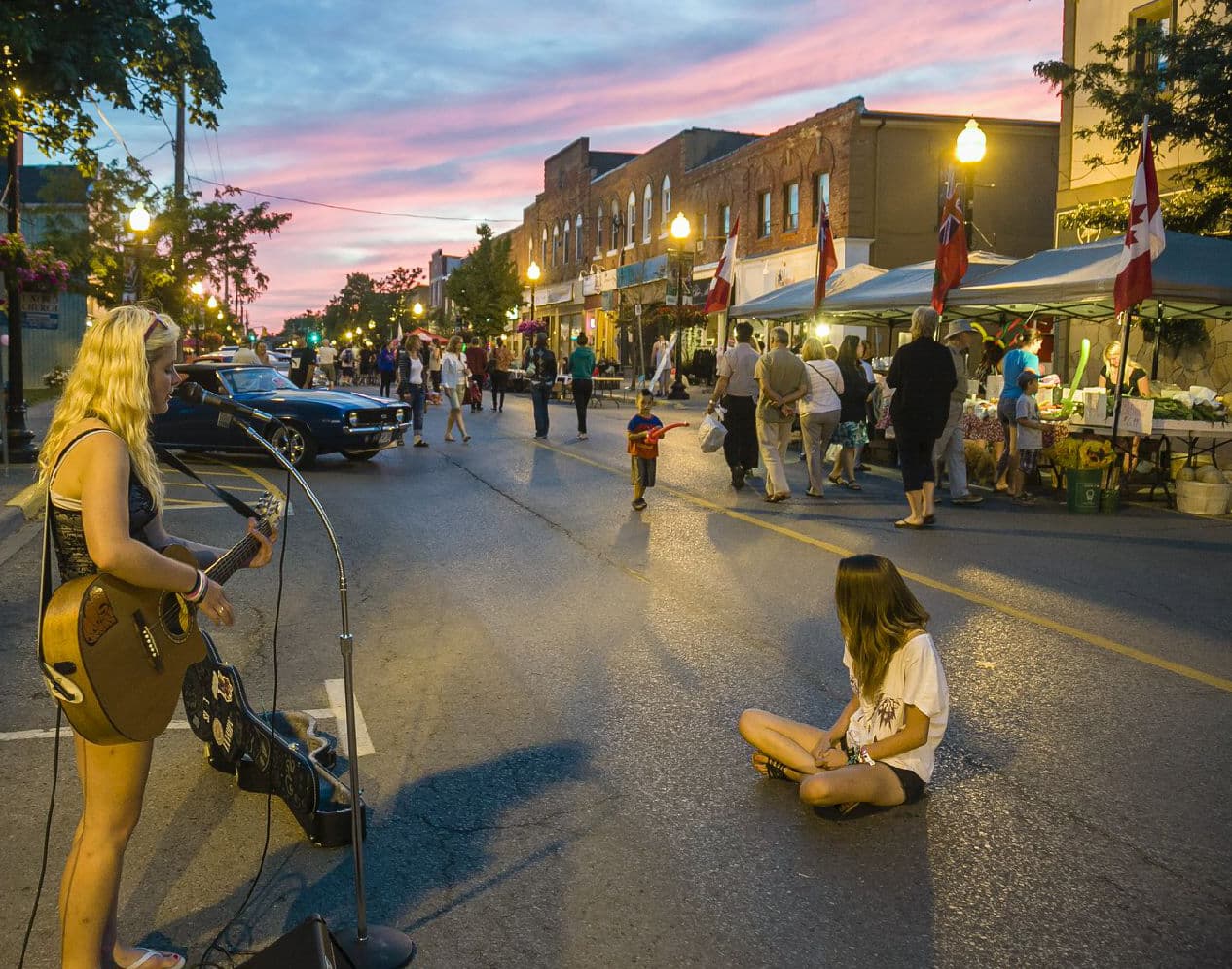 A summer-evening street festival on Main Street Stouffville with a live musician, farmers' market tents and a crowd under a pink sunset